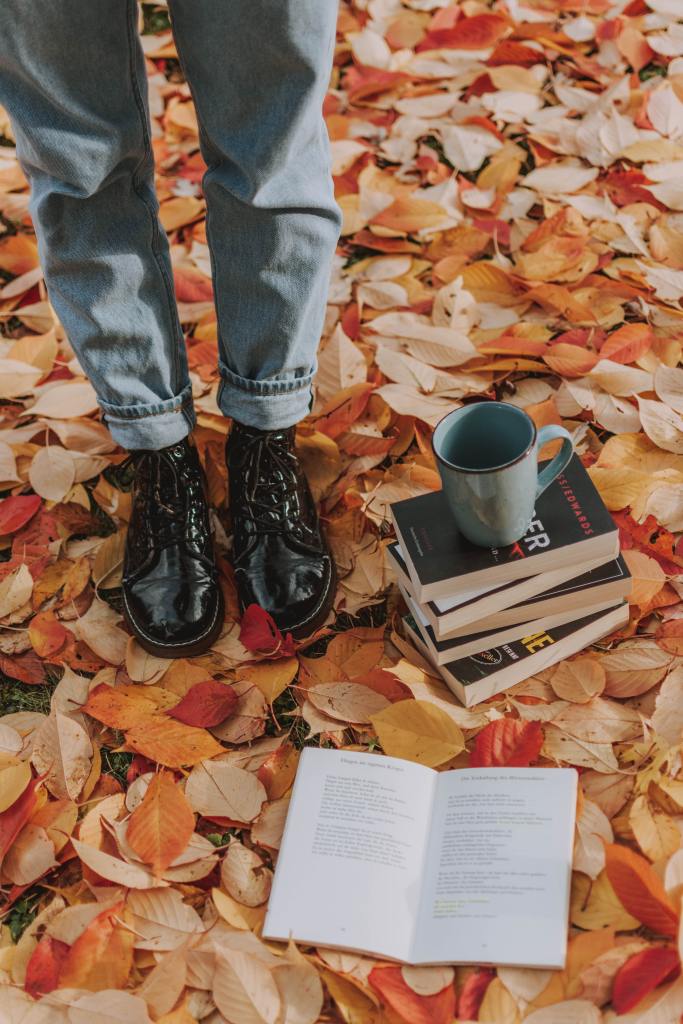 A person standing on fall leaves near a stack of books and a cup of coffee