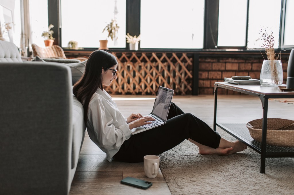 A young woman sitting on the floor of her apartment working on a laptop